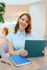 Portrait of a confident businesswoman sitting at a table in a modern office or coworking space. She is smiling warmly and looking at the camera while holding an open notebook. 