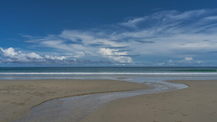 The stream winds along the sandy beach, flows into the ocean. Balls of sand bubbler crabs are visible . The waves are foaming. Blue sky, clouds. Malaysia. Borneo. Kota Kinabalu.