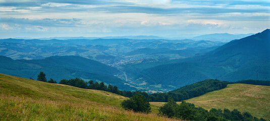 Fototapeta premium A sweeping panoramic view of verdant mountain ridges and valleys under a clear blue sky, with sunlit fields and distant villages nestled among lush green forests.