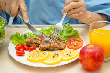 Asian senior woman patient eating pork chop stake and vegetable salad for healthy food in hospital.