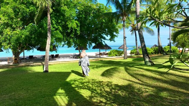 Aerial view of a blonde adult woman in a patterned robe walking from a tropical beach villa toward the sea in Mauritius