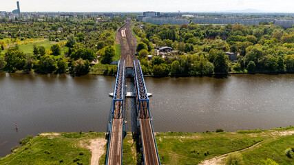 Top view of dual railway bridge tracks extending across a river in lush Polish landscape with dense forest and city in background