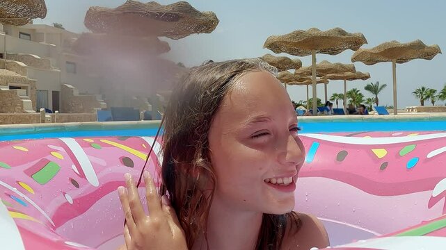Cheerful teenage girl floating on pink inflatable donut ring, splashing and making playful expressions while enjoying sunny resort swimming pool during summer vacation