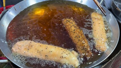 A vendor is frying corndogs in a large pot of hot oil at a bustling night market. The footage captures the sizzling sound and golden-brown color of the corndogs as they cook.