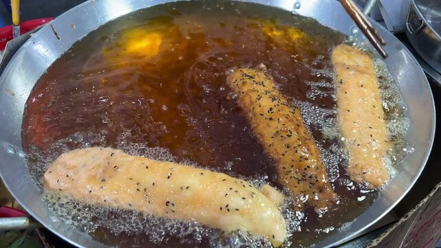 A vendor is frying corndogs in a large pot of hot oil at a bustling night market. The footage captures the sizzling sound and golden-brown color of the corndogs as they cook.