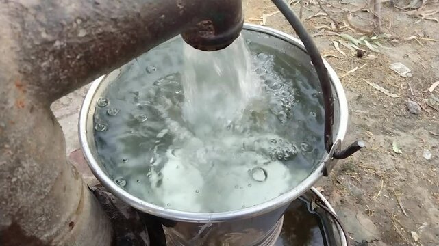 Water flowing from a hand pump into a metal bucket in a rural outdoor setting, symbolizing traditional water access

