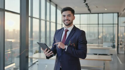 A confident businessman in a suit holding a tablet stands in a modern office with large windows and a city view. - Powered by Adobe
