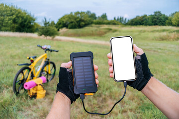 Close-up mockup of a smartphone with a Power Bank charger with a solar panel in the hands of a man. Background of green grass bicycle in nature.