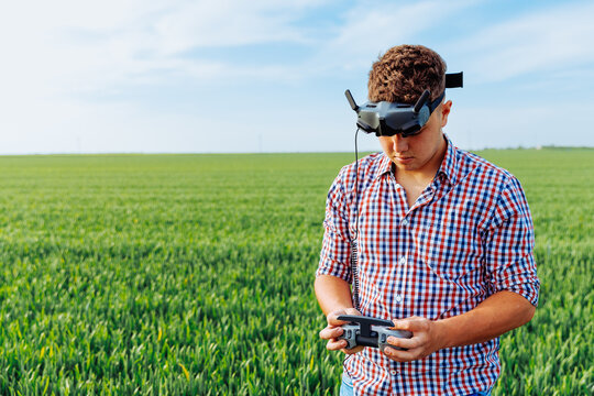 Curly hair young man wears plaid shirt and VR headset while operating drone in green crop field under blue sky