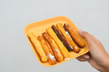 closeup of curos on a yellow plate held by a woman's hand against a white background, is a food made from fried pastry dough, usually choux pastry.