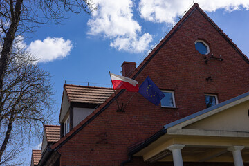 Polish and European Union flags waving side by side on traditional red brick building under blue...