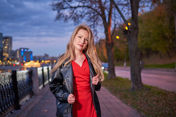 Fototapeta premium Woman in red dress is walking along embankment. She is wearing black jacket. Action takes place at night, with cityscape in background.