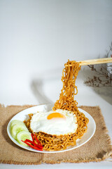 Delicious Indonesian fried noodles served with a sunny side up egg, cucumber slices, and chili, styled beautifully with chopsticks lifting the noodles. Perfect for food blogs or menus.