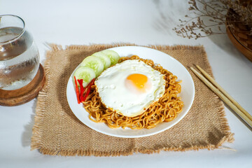 A delicious plate of Indonesian fried noodles topped with a sunny side up egg, served with cucumber slices and chili. Styled with chopsticks and a glass of water for a fresh, homey vibe.