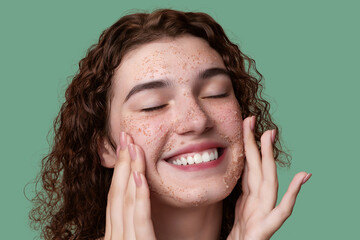 Beauty portrait of a young girl with curly hair joyfully massaging a pink exfoliating face mask...