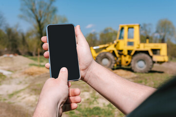 The phone is in the hands of a man. Against the background of a yellow excavator with a wheel...