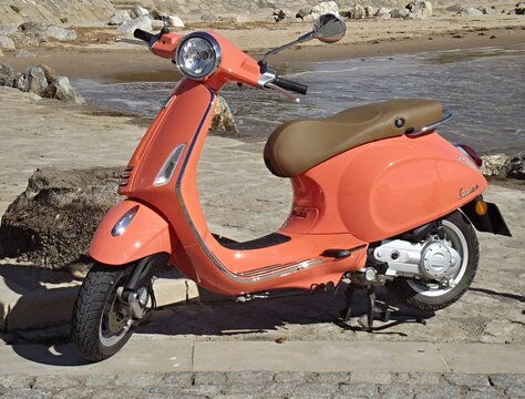 Stylish coral Vespa Primavera 50cc scooter parked on a paved walkway by the beach