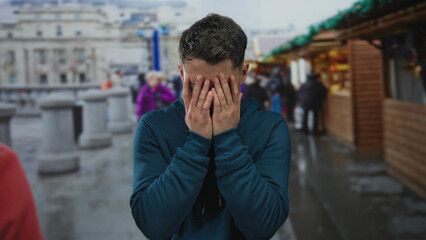 Obraz premium Young hispanic man in market street crying with emotion under cloudy sky, surrounded by bustling store and city life, adorned in casual blue hoodie.