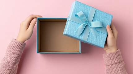 Fototapeta premium Hands are holding a light blue gift box with a ribbon, revealing the inside of the empty container. The pink background contrasts with the box, making the moment visually appealing.