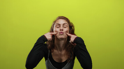 Woman making funny face with fingers on cheeks against a bright yellow background, expressing humor...