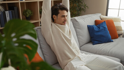 Young man stretching arms in modern living room with cozy sofa and colorful cushions, expressing relaxation in stylish apartment environment, captured during daylight.