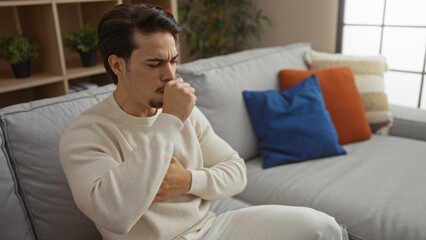 Young man coughing indoors on a cozy sofa with colorful cushions, showcasing a relaxed living room atmosphere with houseplants and soft natural light.
