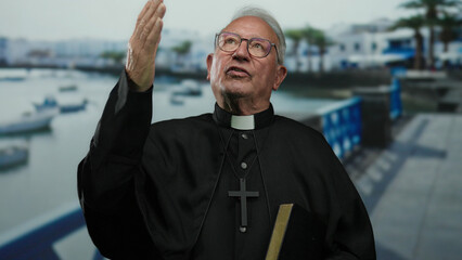 Senior priest man in black robes gestures thoughtfully on a sunny seaside promenade with blue...