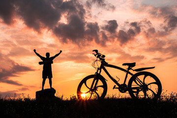 Obraz premium Silhouette of a mountain bike nearby, a happy male cyclist with his arms raised to the top. Sunset background.