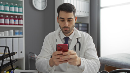 Young hispanic male doctor in hospital room using smartphone with medical equipment and files...