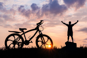 Silhouette of a male cyclist with arms raised top of head. Background sunset sun.