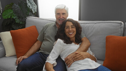 Middle-aged couple sitting closely on a cozy couch smiling, man embracing woman indoors in a...