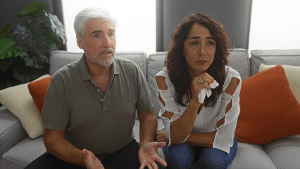 Middle-aged couple sitting in a living room, man and woman appear emotional while discussing,...