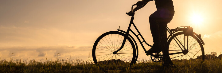 Long Banner. Close-up silhouette of a bicycle with cyclist's legs at sunset. Background of a field...