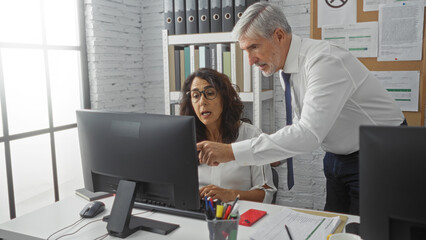Man and woman collaborating at a desk in a bright office, focused on a computer screen with files and documents, showcasing teamwork in a modern workspace setting.