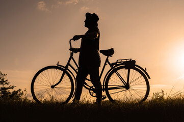 Silhouette of a walking man in a hat next to a bicycle. Sunset background.