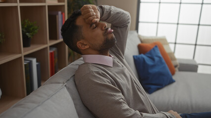Young hispanic man sitting indoors in a stylish apartment living room looking contemplative and...