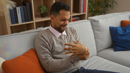 Fototapeta premium Young man sitting on a sofa indoors experiencing hand discomfort, surrounded by colorful cushions and shelves in a cozy, contemporary living room setting.
