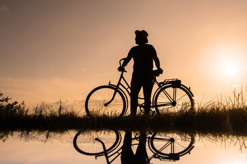 Silhouette of a cyclist with water reflection. Sunset background.