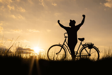 Silhouette of a cyclist with a bicycle with his arms raised on top at sunset with the symbol of...