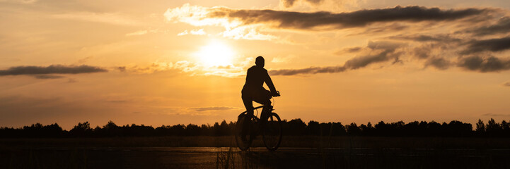 Long Banner. Close-up silhouette of a bicycle with cyclist's legs at sunset. Background of a field...