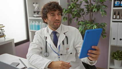 Fototapeta premium Handsome hispanic male doctor in a white coat using a tablet indoors in a clinic room, surrounded by medical equipment and greenery indicating a healthcare setting.