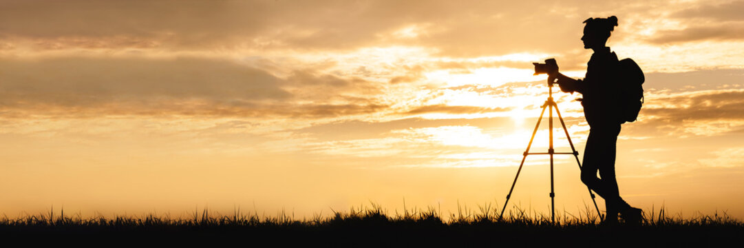 Long banner silhouette of a female photographer with a photo-video camera on a tripod, standing in a meadow in a field. Background of sunset on sky with clouds. - Powered by Adobe