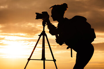 Silhouette of a young female photographer with a professional photo video camera on a tripod....