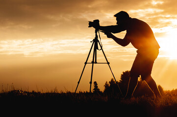 Silhouette of a photo-video camera standing on a tripod with a male photographer filming in a...
