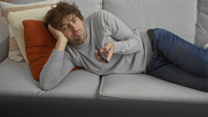 Fototapeta premium Man relaxing on a sofa with a remote control in a cozy living room appearing thoughtful and focused.