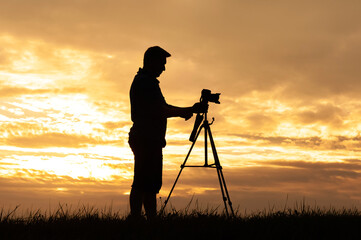 Silhouette of a photographer with a professional camera standing on a tripod. Against the background of the sunset sky.
