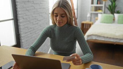 Woman working on laptop in cozy bedroom setting holding card, suggesting online shopping or work...