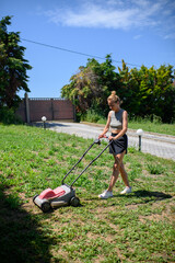 Young woman mowing green grass with a lawn mower on a sunny day near a cozy countryside house. Summer gardening work, rural lifestyle and outdoor chores concept.