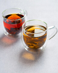 Two cups of assorted black and green tea in transparent double glassware on light grey background.