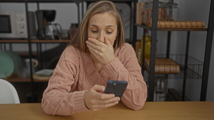 Woman in office looks at smartphone with surprise, hand covering mouth, sitting indoors in a...